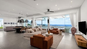 A spacious living area with a large sofa, a coffee table, and a TV. There is a kitchen area with a dining table and chairs, and a view of the ocean through large windows. - Golden Lotus Villas (Koh Samui)