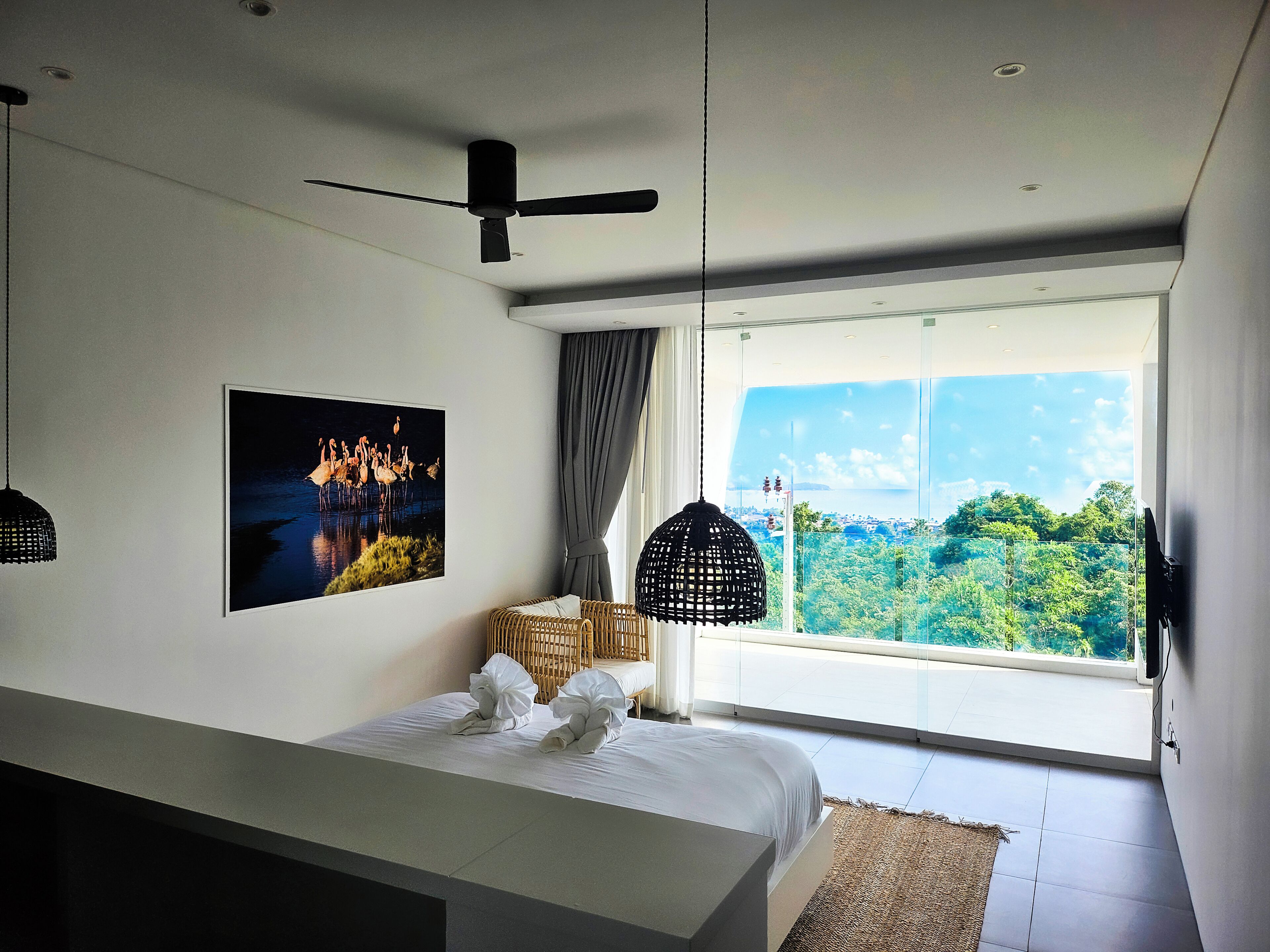 Un dormitorio moderno con un ventanal grande, un ventilador de techo, una cama con ropa blanca, un televisor montado en la pared y un cuadro con flamencos.