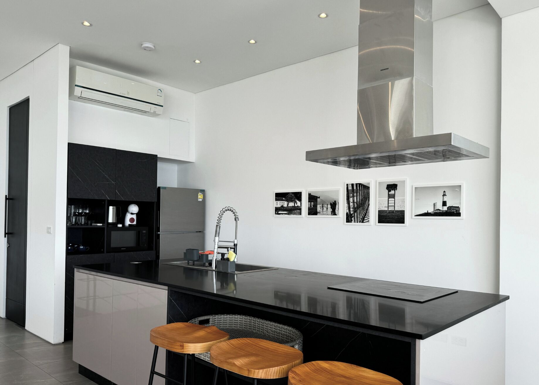A modern kitchen with a central island, bar stools, a stainless steel range hood, and a wall-mounted air conditioner.