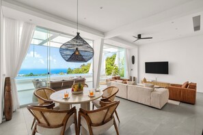 A modern living room with a dining area, large windows, a flat-screen TV, and a view of the ocean. - Golden Lotus Villas (Koh Samui)