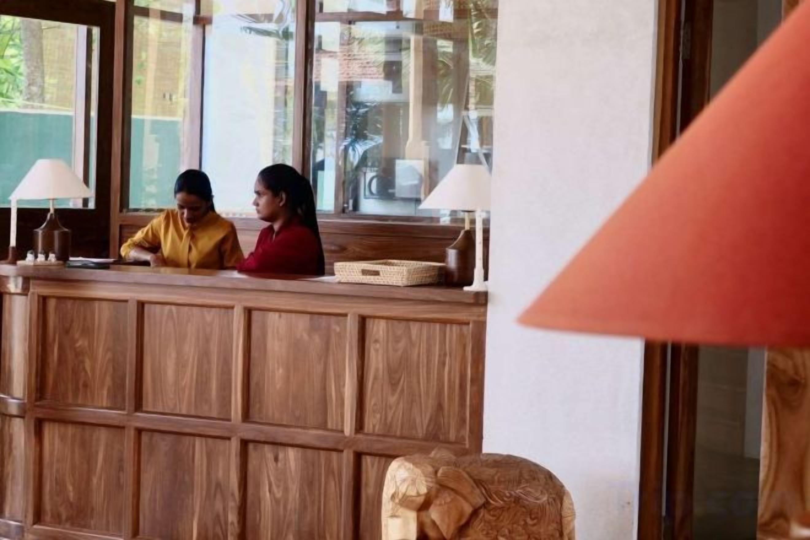 A reception desk with two staff members, a wooden panel design, and a lamp.