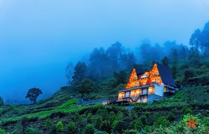 Un bâtiment à deux étages, doté d’un toit en pente, entouré d’une végétation luxuriante et de montagnes brumeuses.
