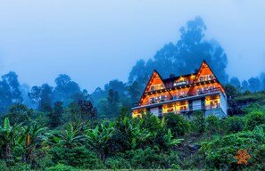 Un chalet à deux étages, doté d’un toit triangulaire, entouré d’une végétation luxuriante et d’arbres.