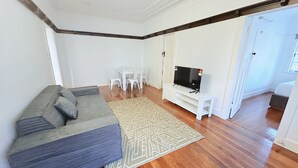 Un salon avec un canapé gris, un tapis à motifs, un téléviseur posé sur un meuble blanc et un coin repas avec des chaises blanches.