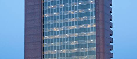 Un edificio alto y moderno con una 'R' destacada en el lado izquierdo, con fachada de vidrio y base de ladrillo.