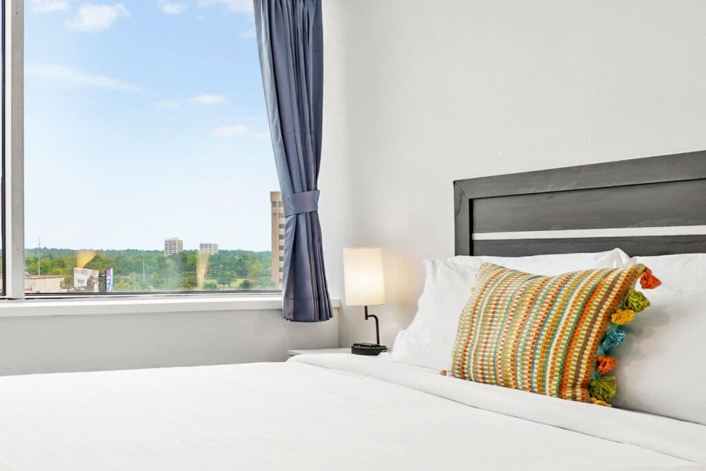 A hotel room with a bed, a window with curtains, a nightstand with a lamp, and a view of buildings outside.