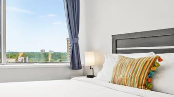 A hotel room with a bed, a window with curtains, a nightstand with a lamp, and a view of buildings outside.