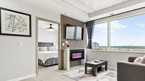 A modern living room with a fireplace, a sofa, a TV mounted on the wall, and a large window with a city view. - Reese Tower (Tulsa)