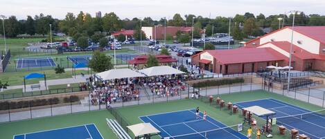 Complejo de canchas de tenis con varias canchas, asientos para espectadores y un edificio de techo rojo.