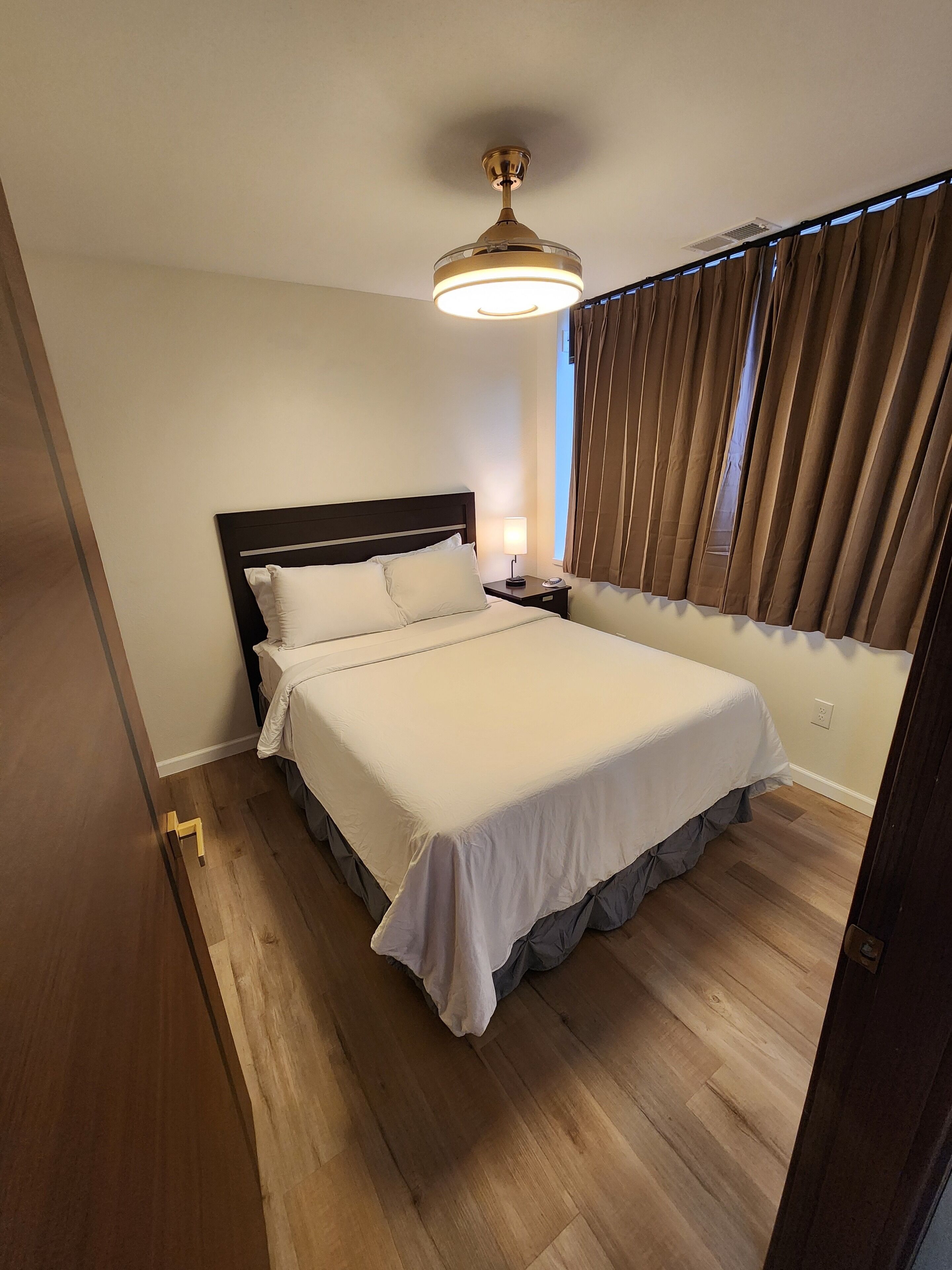Un dormitorio con una cama grande, una mesita de noche, un ventilador de techo y una ventana con cortinas.
