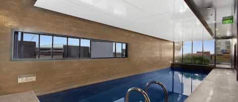 An indoor swimming pool with clear glass walls, a visible exit sign, and a window with a view of the outside.