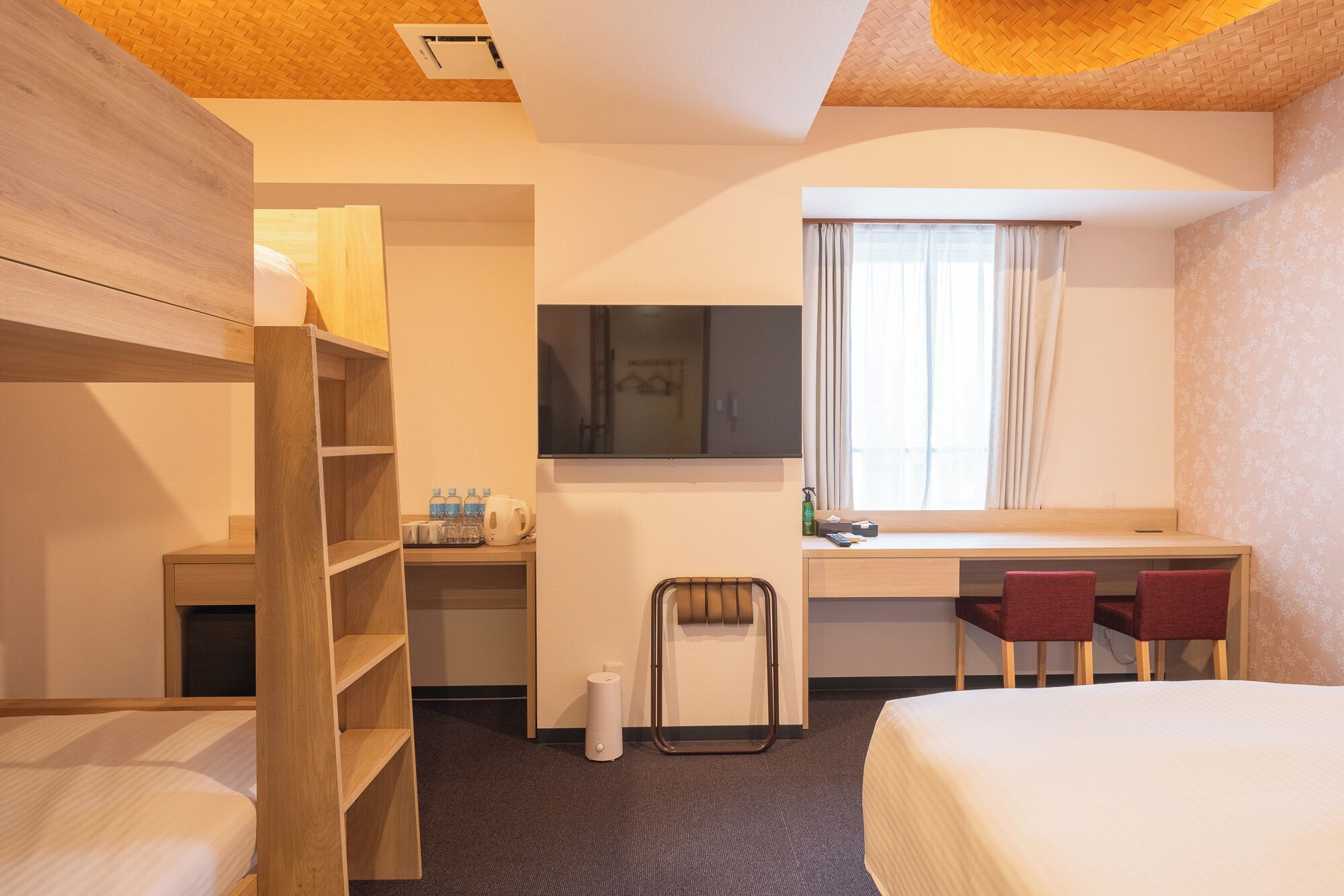 A hotel room with a bed, a desk with two chairs, a TV, a window with curtains, a shelf with toiletries, and a wooden wardrobe.