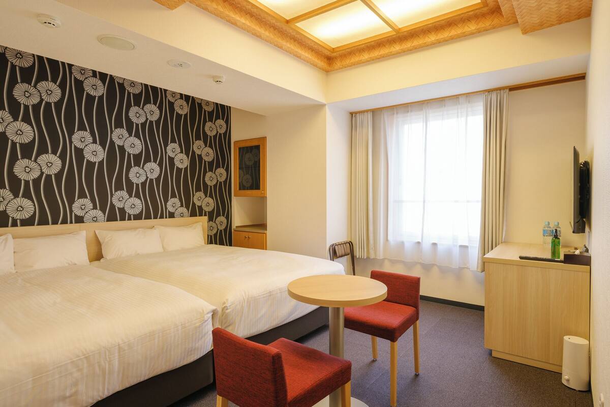 Een hotelkamer met twee bedden, een klein rond tafeltje, twee rode stoelen, een bureau met een spiegel en een raam met gordijnen.