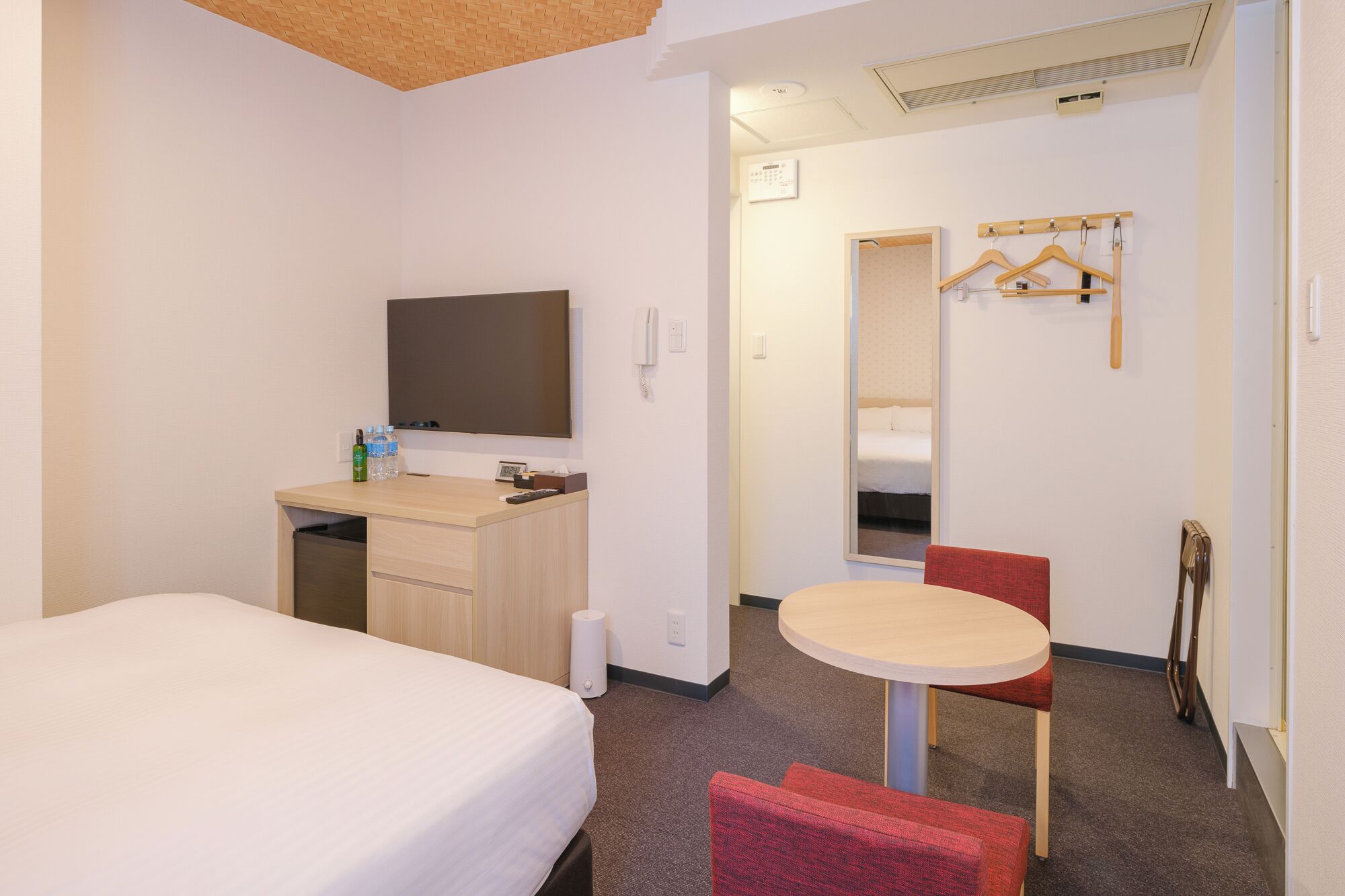 A hotel room with a bed, a desk with a flat-screen TV, a small table with a chair, a mirror, and a coat rack with hangers.