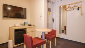 Una habitación de hotel con un escritorio de madera, dos sillas rojas, una mesa redonda, un espejo, un perchero y un pequeño cubo de basura.