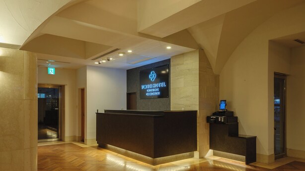 Zona de recepción de un hotel con diseño moderno, que incluye un mostrador de recepción, una pantalla digital con el nombre del hotel y un suelo de madera con patrón de espiga.