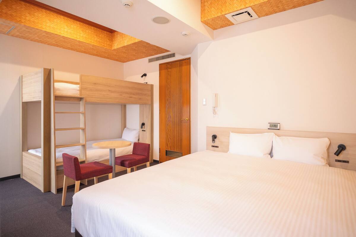 Een hotelkamer met een stapelbed, een eenpersoonsbed, een bureau met stoel, een telefoon en een wekker.