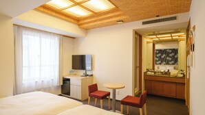 Habitación de hotel con dos camas, un escritorio, televisor, una mesita y baño con espejo y lavamanos.