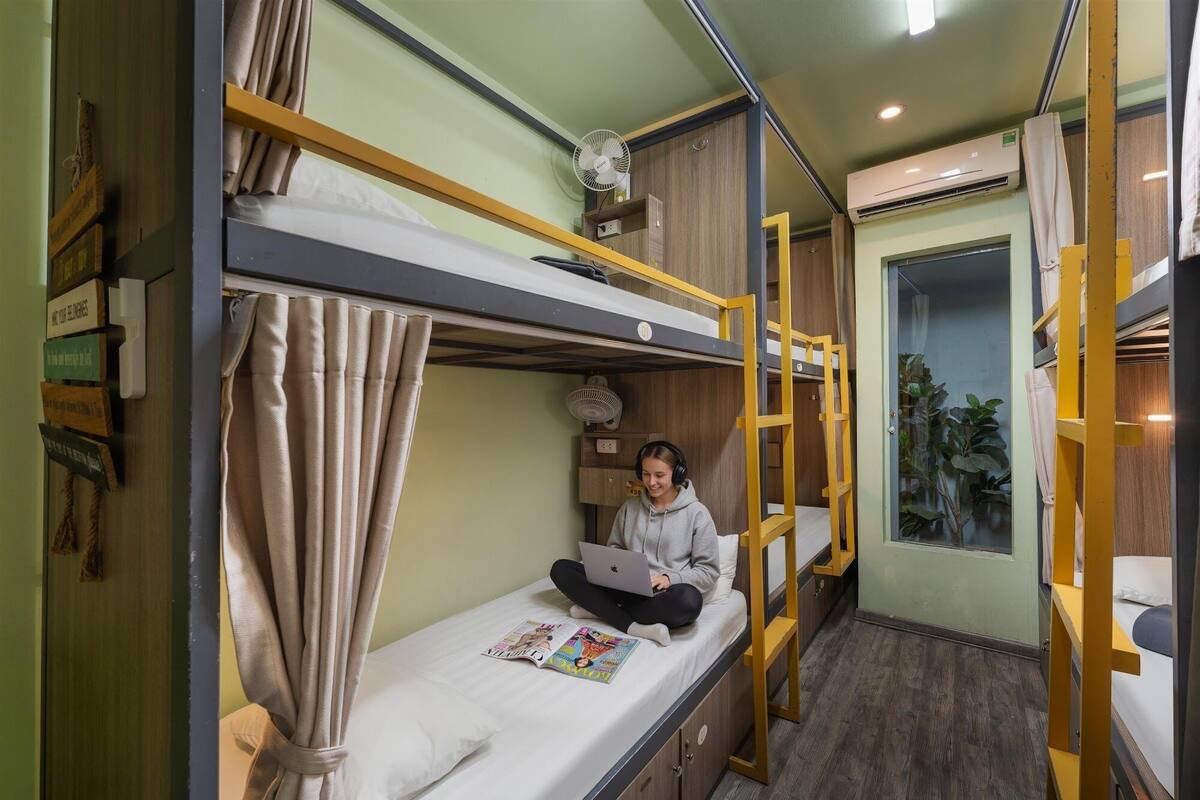 Habitació amb lliteres, un ordinador portàtil al llit inferior, un ventilador al llit superior i una finestra amb cortines.