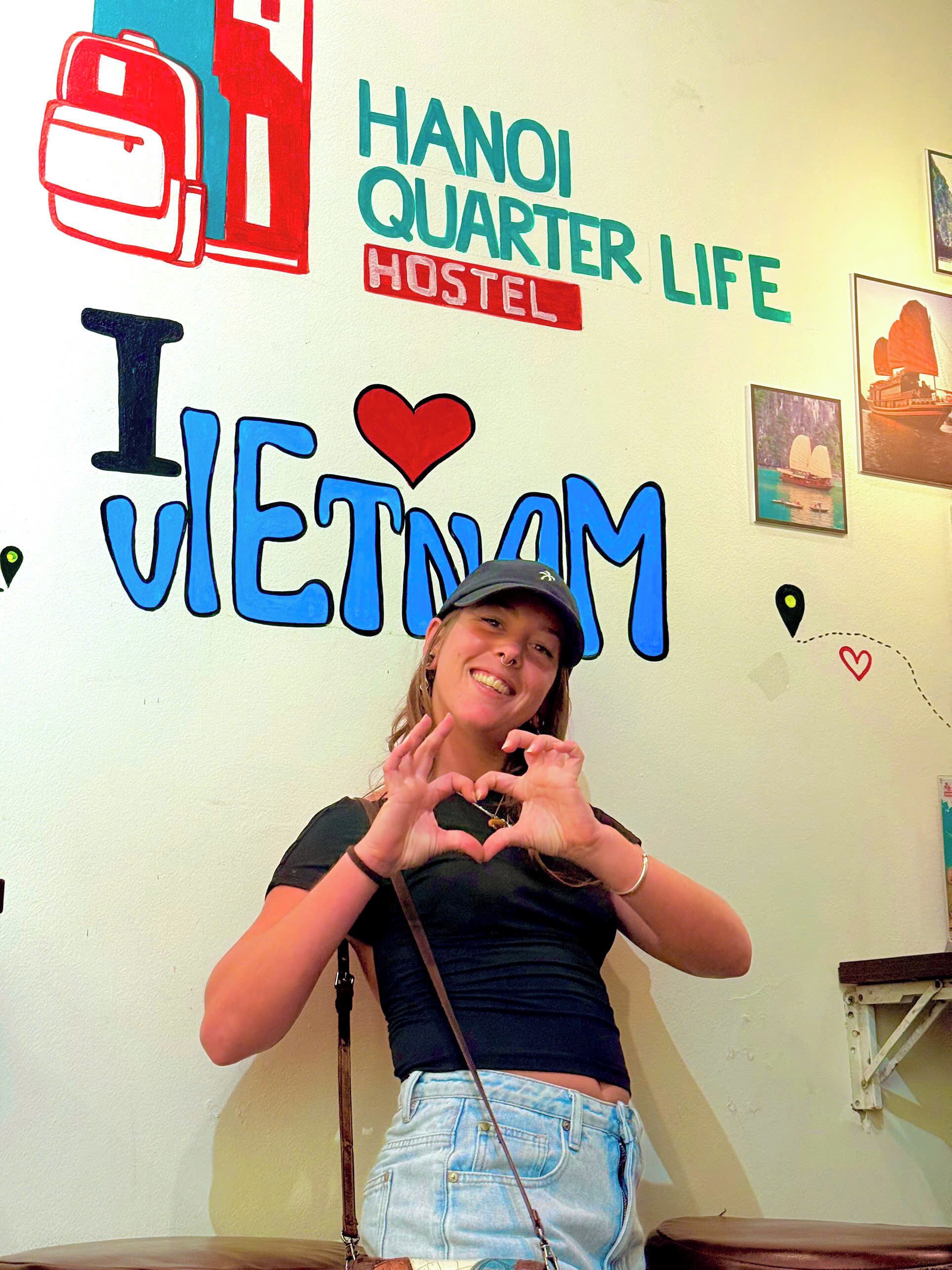 Una pared decorada con 'HANOI QUARTER LIFE HOSTEL' y 'I ♥ VIETNAM', una persona formando un corazón con las manos y una mochila.