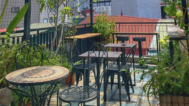 A covered outdoor seating area with tables and chairs, surrounded by potted plants and a view of buildings in the background. - Hanoi Quarter Life Hostel (Hanoi)