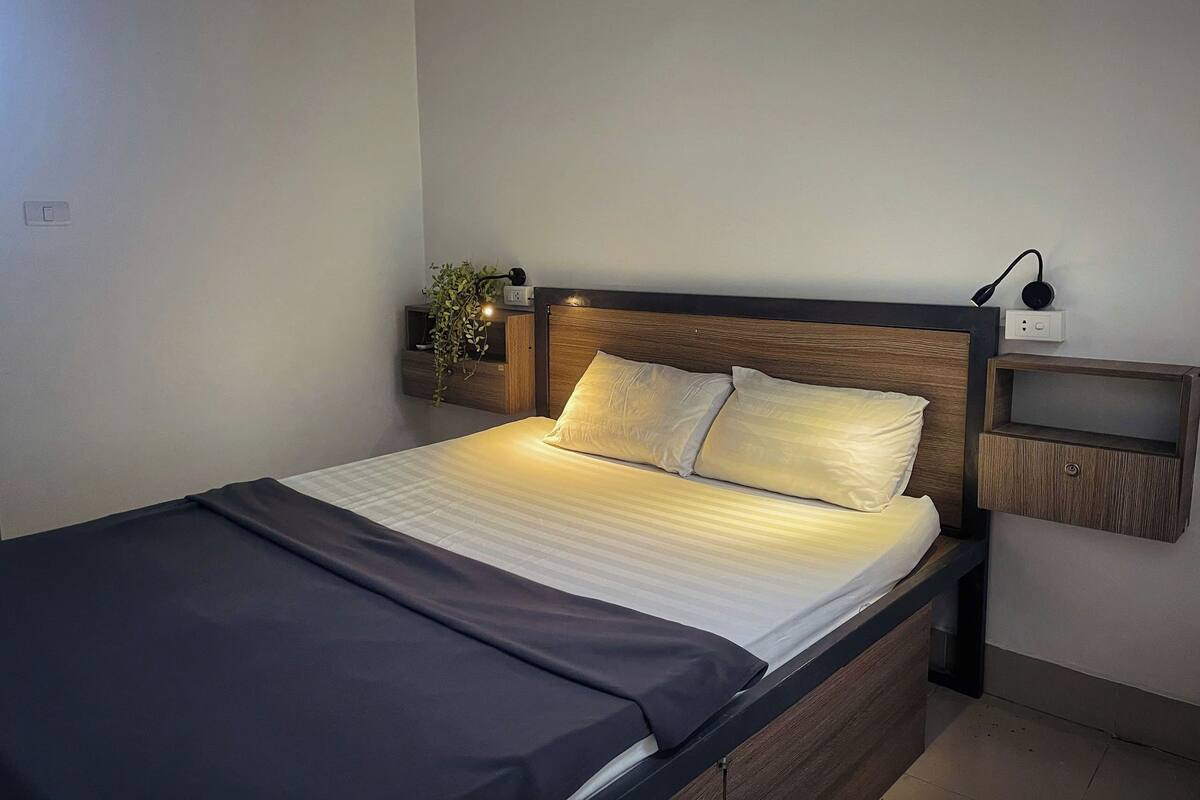 Un llit ben fet amb capçalera de fusta, una taula de nit amb una làmpada i una presa elèctrica penjada a la paret.
