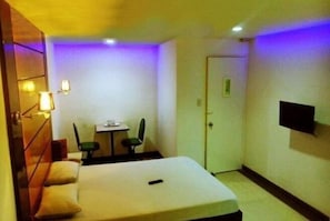 A small hotel room with a bed, a desk, two chairs, a TV, and a door. - Halina Drive Inn Hotel - Pasay (Pasay)