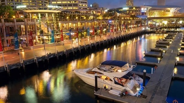 Un puerto deportivo con barcos amarrados, un paseo marítimo y edificios al fondo.