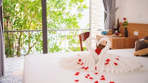 A hotel room with a bed, a wooden side table with a bottle, a vase with a rose, and a view of greenery through the window. - Tam Coc Little Charm Villa & Bungalow (Nam Hoa Lu)