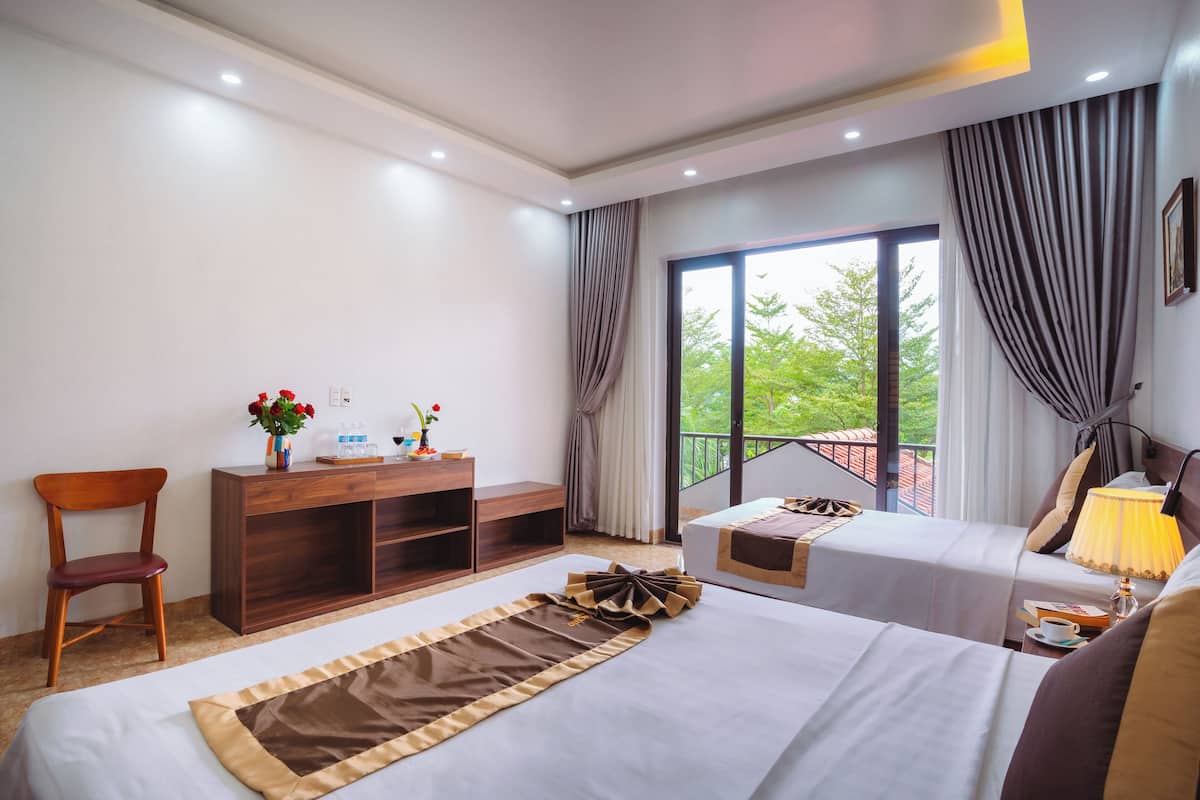 A hotel room with two beds, a desk, a chair, and a balcony with a view of greenery.