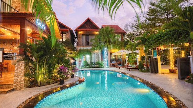 A swimming pool with a tiled edge, surrounded by lush greenery and a building with a red roof. - Tam Coc Little Charm Villa & Bungalow (Nam Hoa Lu)