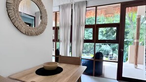 A room with a wooden table, a woven chair, a mirror, and a view through a glass door to an outdoor area. - Villa Bohème, Villa 28 (Koh Samui)