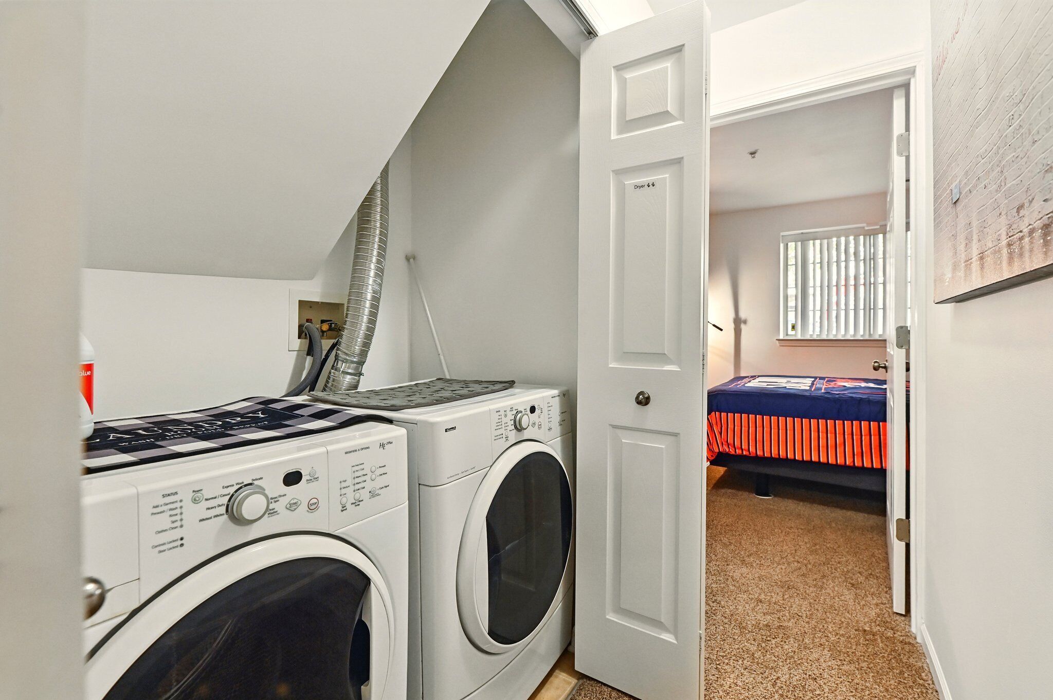 A laundry room with two washing machines, a dryer, and a door leading to a bedroom with a bed and a window.