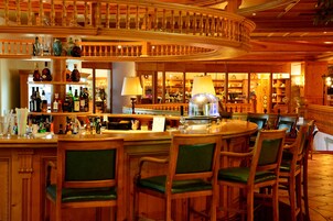A bar area with wooden furnishings, a counter with various bottles, and a seating arrangement with green upholstered chairs.
