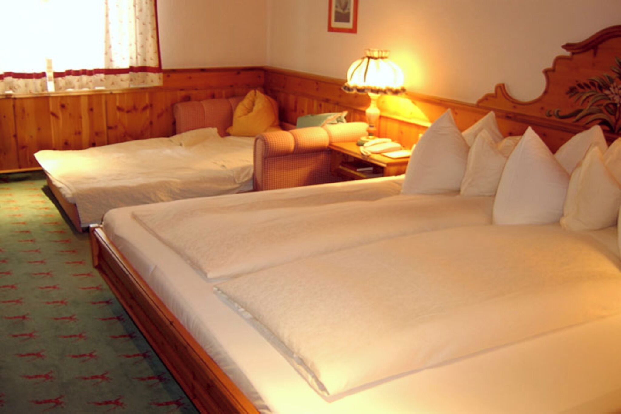 Habitación de hotel con dos camas individuales, cabecera de madera, mesita de noche con lámpara y alfombra estampada.