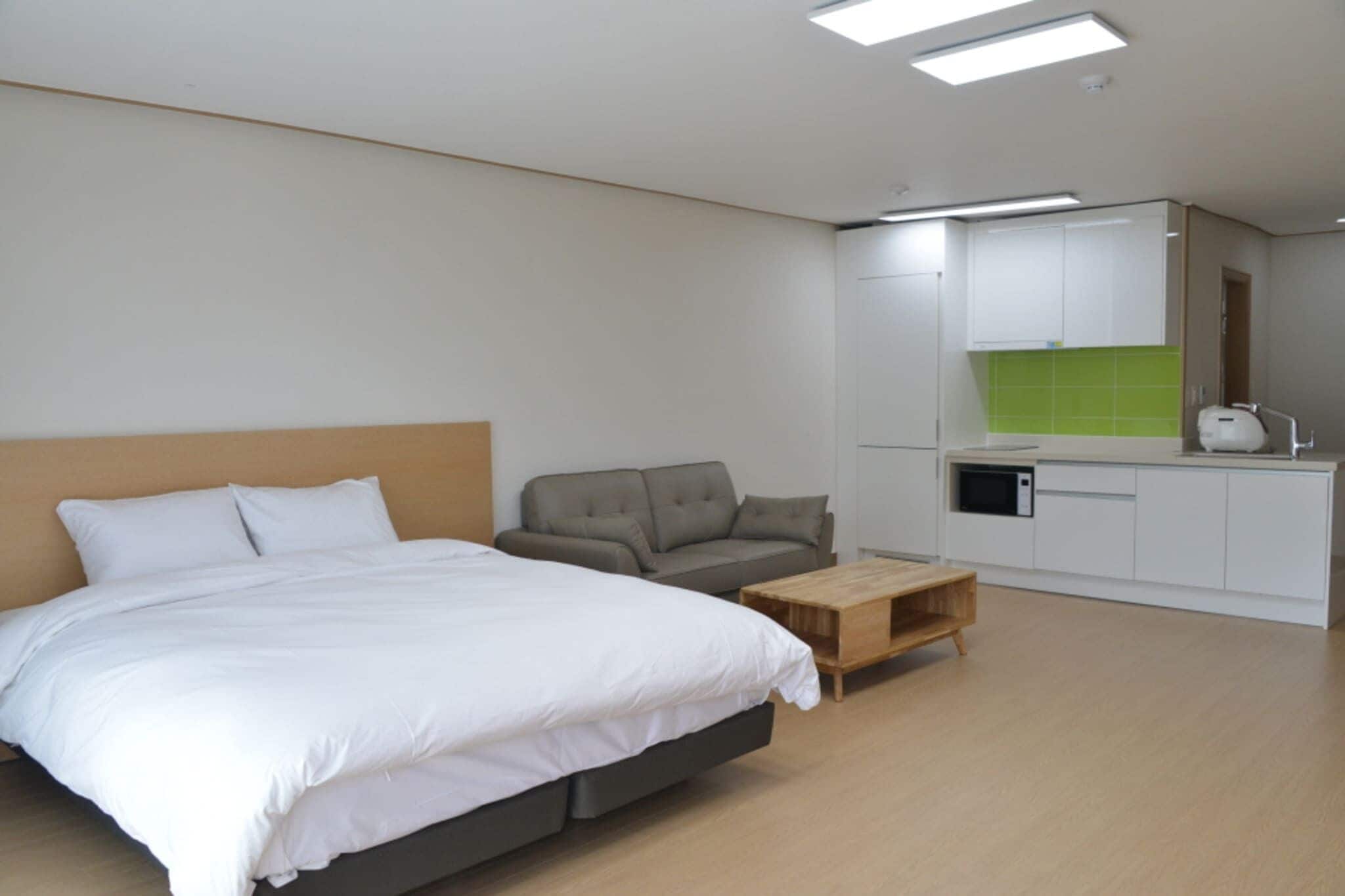 A modern hotel room with a large bed, a sofa, a wooden coffee table, and a kitchenette area.