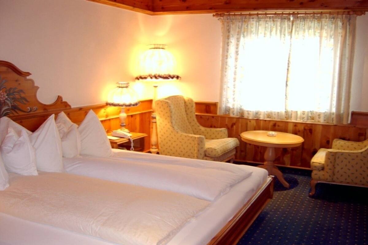 Une chambre d’hôtel avec un grand lit, deux fauteuils, une petite table ronde et une fenêtre avec des rideaux.