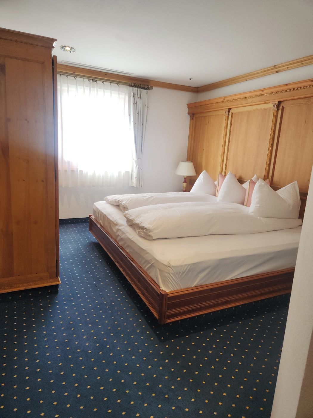 A hotel room with a large bed, a wardrobe, a window with curtains, and a patterned carpet.