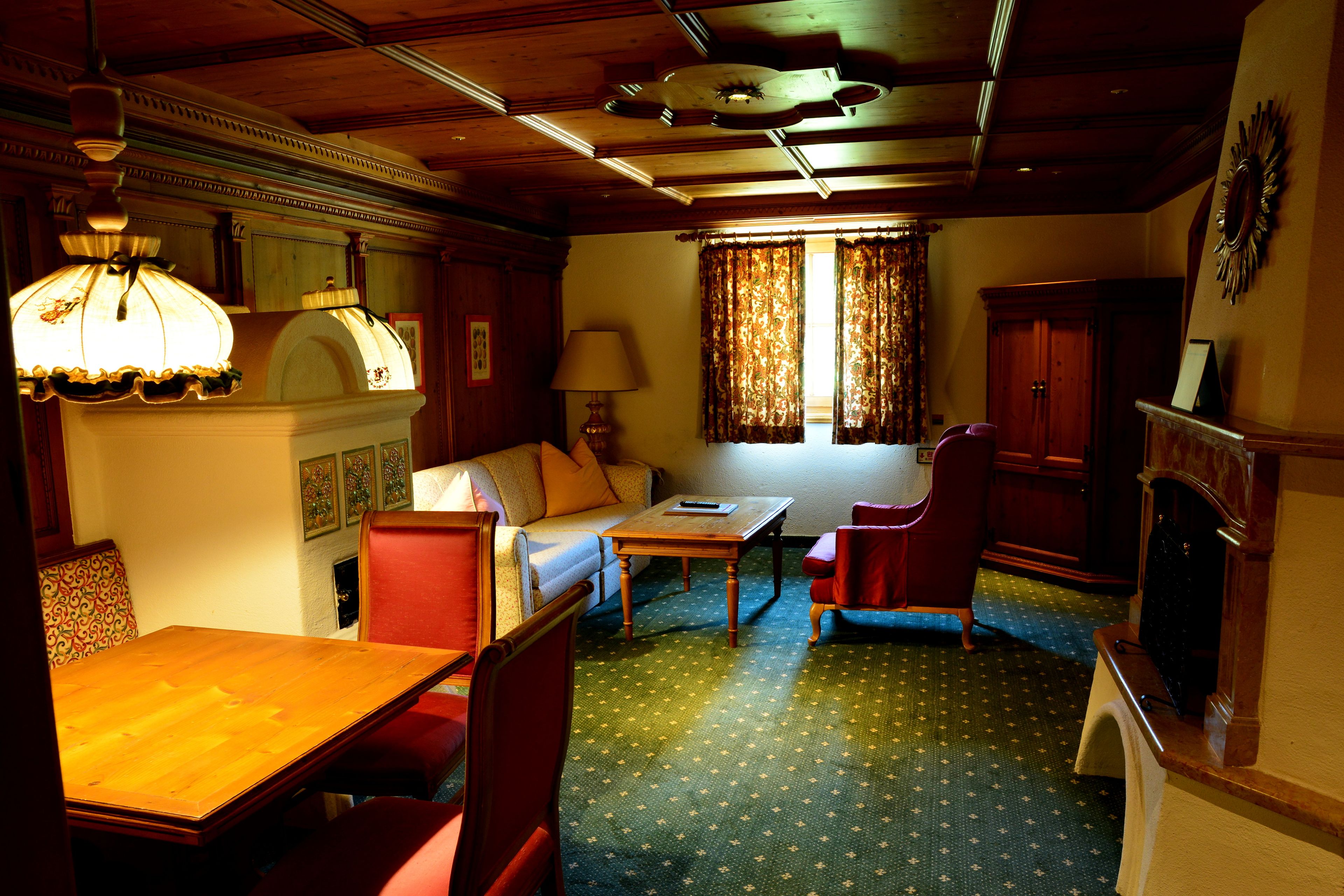 A room with a wooden ceiling, a sofa, a table, a chair, a lamp, and a window with curtains.