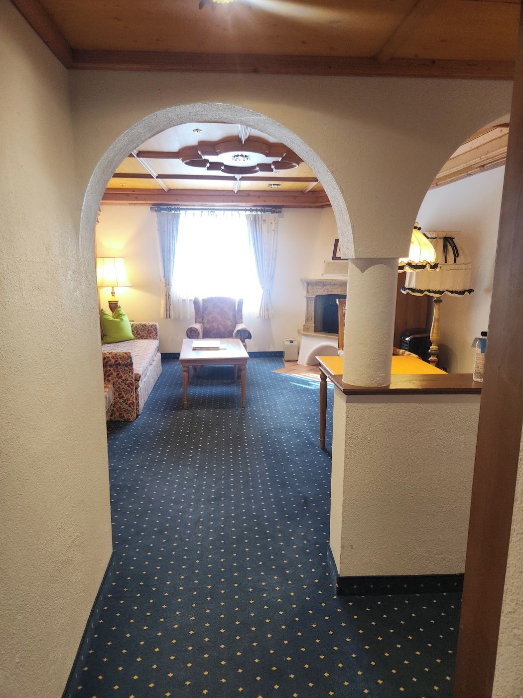 A hotel room with a blue carpet, a wooden table, a sofa, and a lamp.