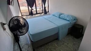 A small, compact room with a single bed, a standing fan, and a window with curtains. - Hospedaje ASL HOME (Yumbo)