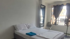 A hotel room with a bed, a chair, a mirror, a window with curtains, and a standing fan. - Hospedaje ASL HOME (Yumbo)