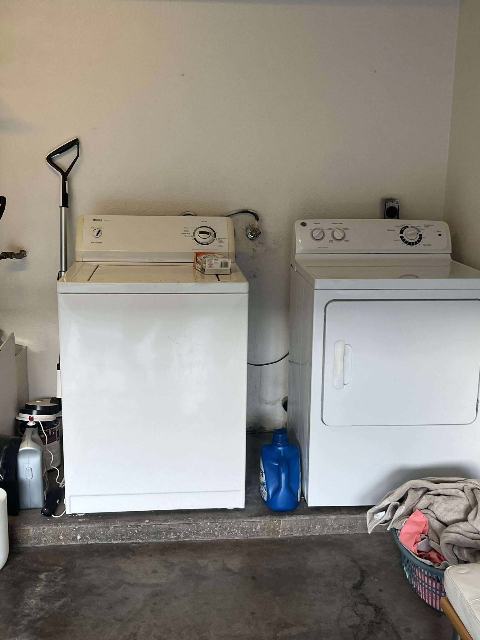 A laundry area with two white washing machines, a blue detergent bottle, and a basket of clothes.