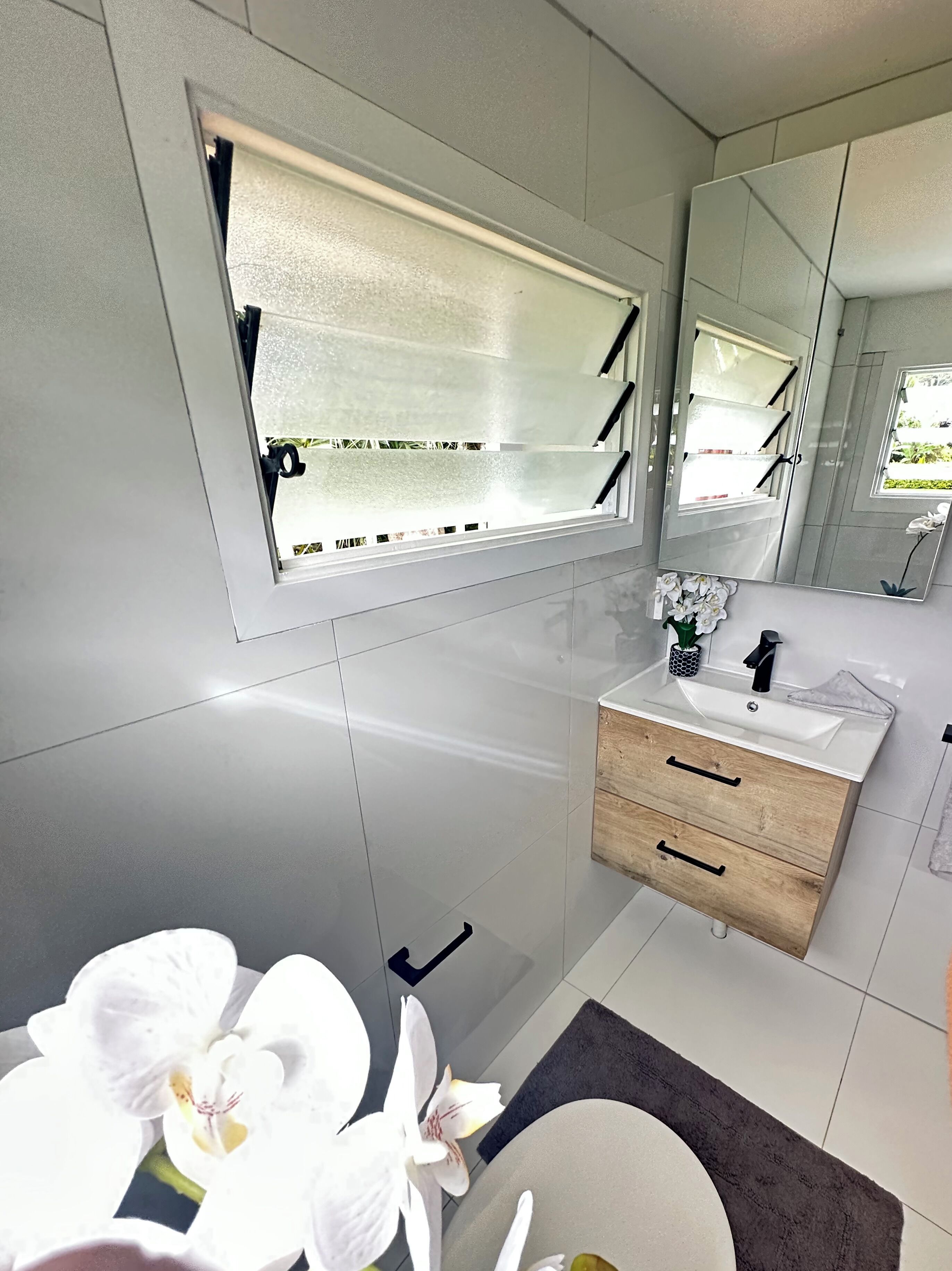 A modern bathroom with a large mirror, a window with blinds, a sink, and a wooden cabinet.