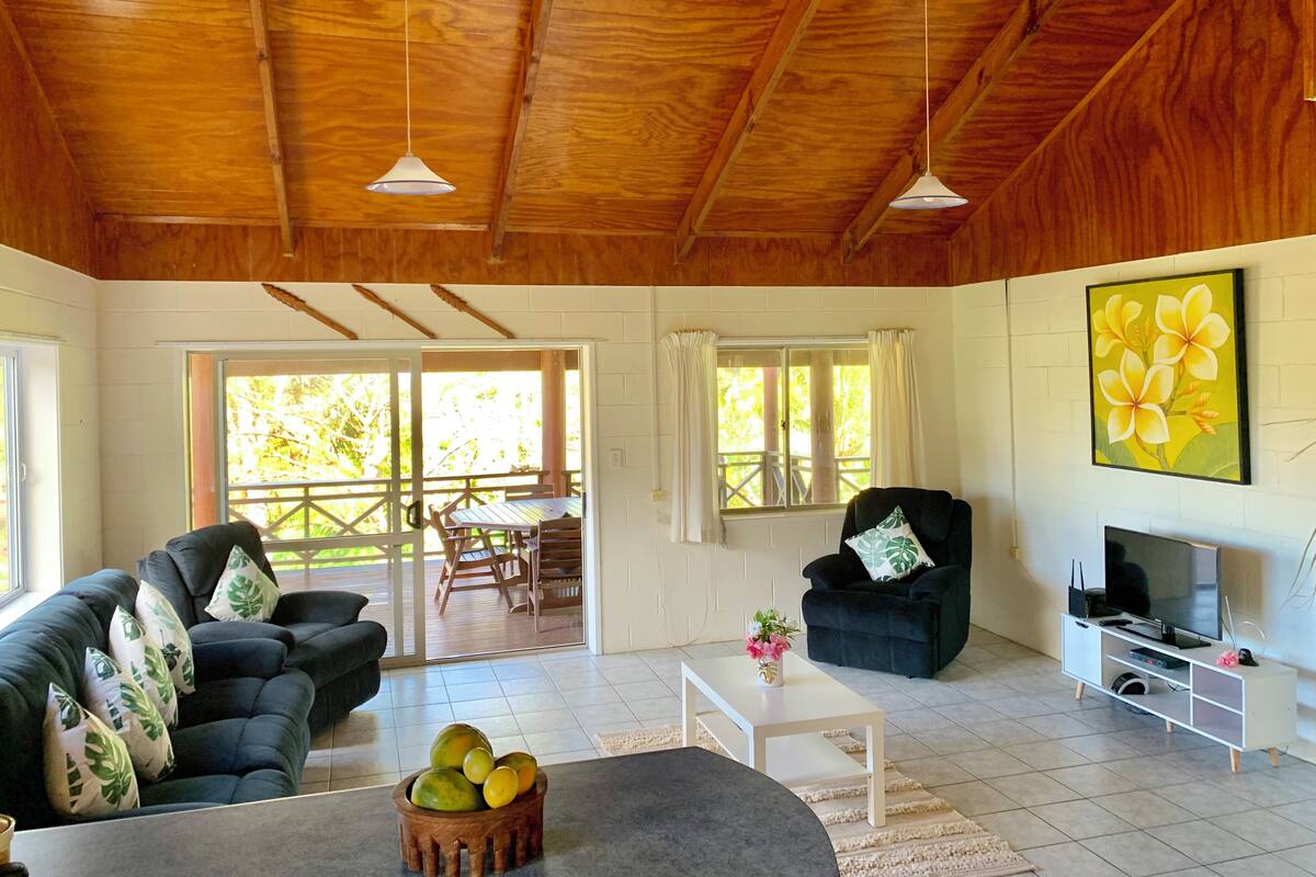 A spacious living room with a wooden ceiling, a black sofa, a white coffee table, and a television on a white stand.