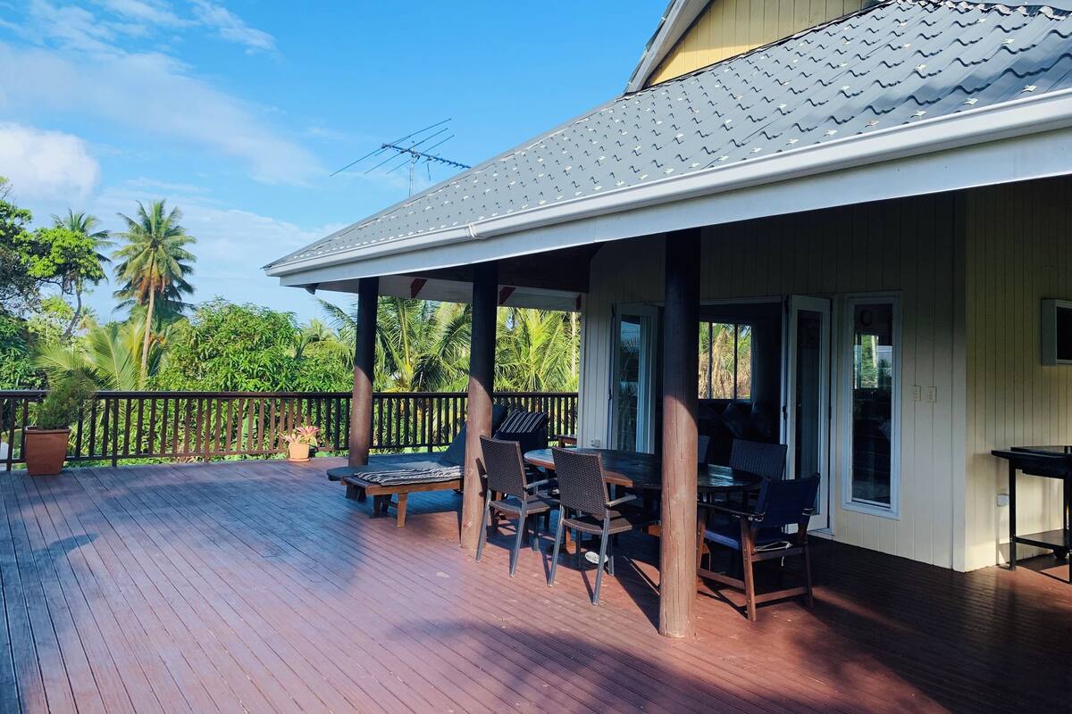 A covered outdoor deck with a dining area, a pergola, and a view of palm trees.