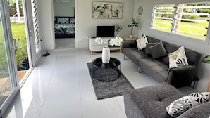 Uma sala de estar moderna com um sofá cinzento, uma mesa de centro e uma televisão. Há uma porta de vidro deslizante que dá para uma varanda com plantas.