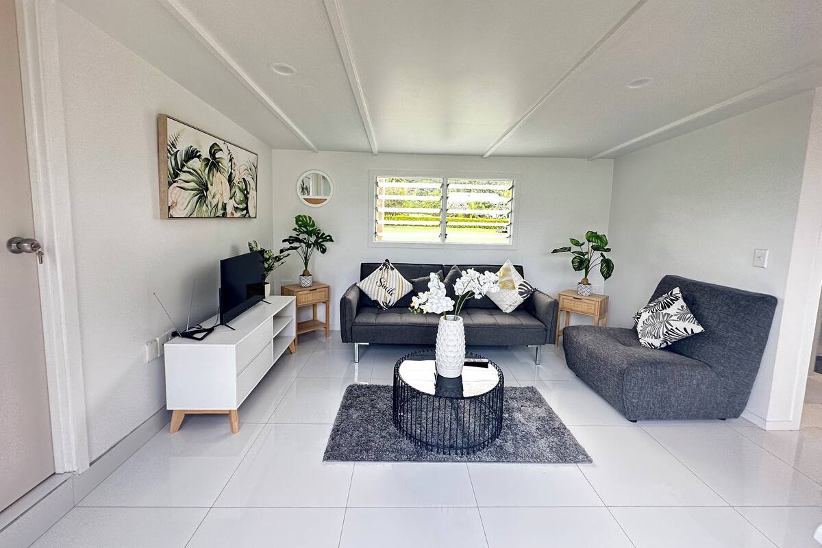A modern living room with a grey sofa, a white TV stand, and a coffee table. There are two windows with blinds, a door, and a painting on the wall.