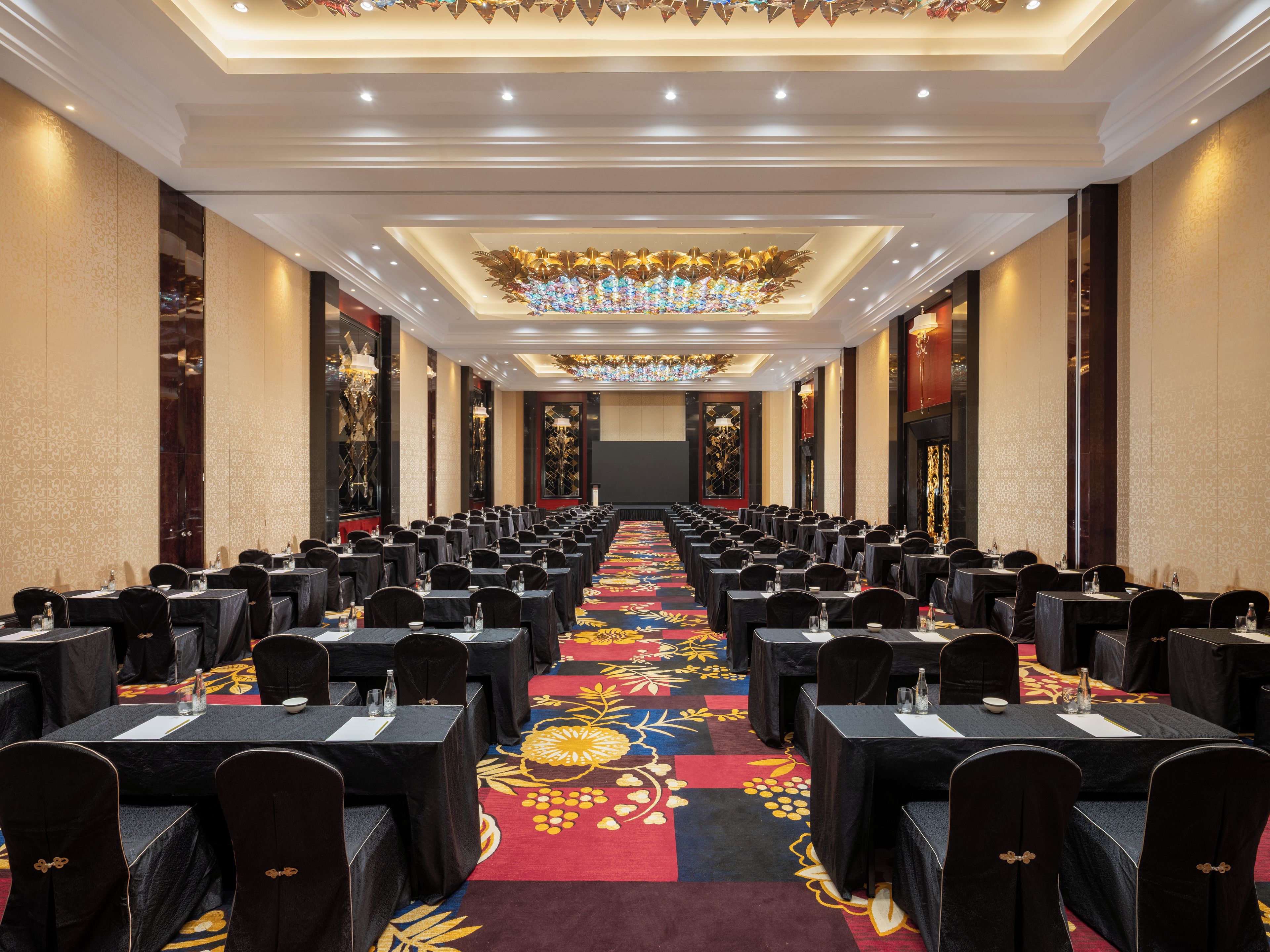 A conference room with rows of tables and chairs, a patterned carpet, and a decorative ceiling light fixture.