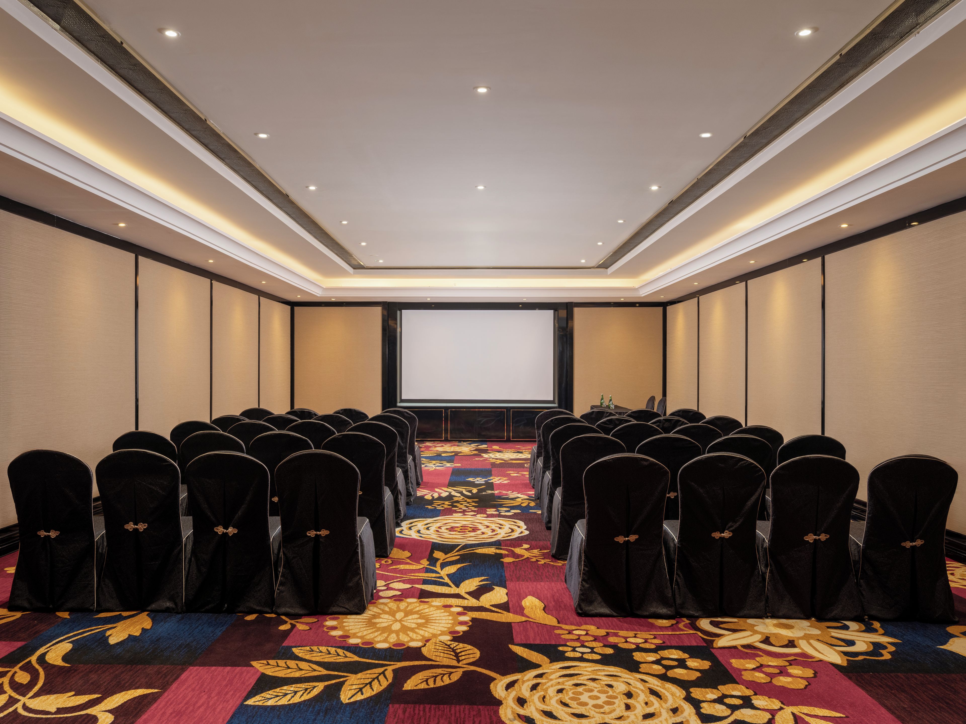 A conference room with rows of chairs, a carpeted floor, a projector screen, and a table with water bottles.